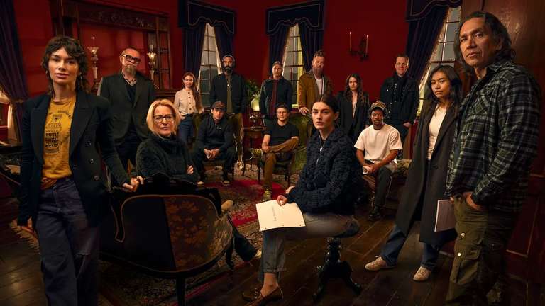 Eine Gruppe von dreizehn Darstellern und Crewmitgliedern posiert auf einem gedämpft beleuchteten, rot getönten Set. Eine Frau hält ein Skript.