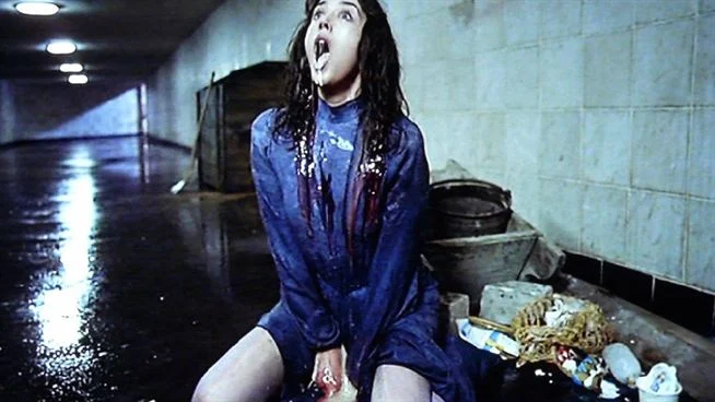 Eine Frau im blauen Kleid kniet schreiend in einem gefliesten U-Bahn-Tunnel, dunkle Flüssigkeit an ihrem Hals.