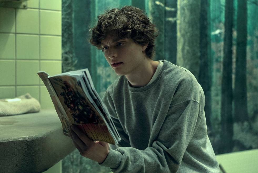 Ein junger Mann mit lockigen Haaren sitzt in einem grün gefliesten Raum und liest konzentriert ein Comic-Heft.