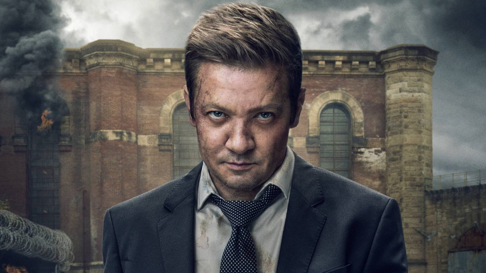 Nahaufnahme eines Mannes, gespielt von **Jeremy Renner**, mit ernstem und intensivem Blick direkt in die Kamera. Er hat kurze, braune Haare und sein Gesicht ist mit Schmutz und Kratzern bedeckt. Er trägt einen dunklen Anzug, ein weißes Hemd und eine gepunktete Krawatte. Im Hintergrund ist ein großes, altes, braunes Ziegelsteingebäude zu sehen, das teilweise von dicken Rauchwolken umgeben ist, die in den grauen, bedeckten Himmel aufsteigen. Eine Flamme ist an der Seite des Gebäudes zu sehen.