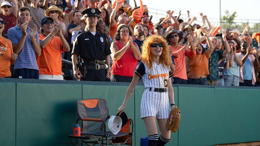 Frau mit roten Locken in Baseballuniform ("Cheesewrong") lächelt mit Handschuh und Mütze vor jubelnder Stadionmenge.