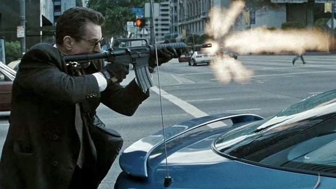 Eine Filmszene auf einer Stadtstraße bei Tageslicht zeigt einen Mann in einem dunklen Anzug und einer Sonnenbrille, der hinter einem blauen Auto in Deckung geht. Er kniet leicht und hält ein Sturmgewehr (wahrscheinlich ein M16 oder AR-15-Derivat) vor sich, wobei Feuer und Rauch aus der Mündung schießen, was auf einen Schuss hinweist. Die Szene ist actiongeladen und dramatisch. Im Hintergrund sind weitere Autos und ein Fußgänger auf der anderen Straßenseite zu sehen, sowie hohe Gebäude.