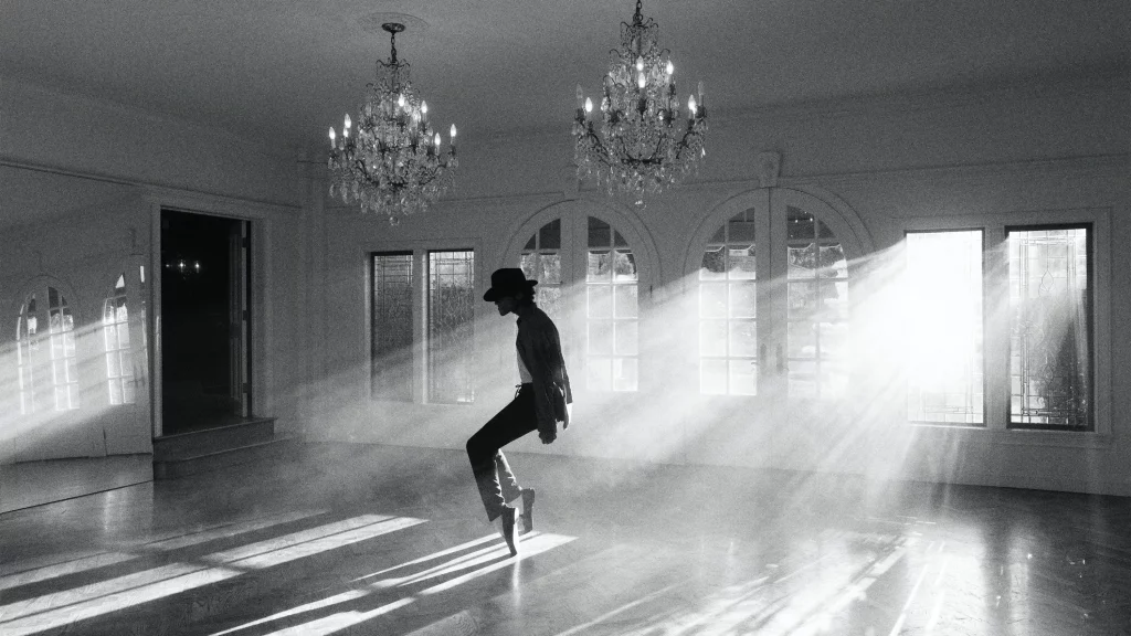 Ein Schwarz-Weiß-Bild zeigt Michael Jackson (oder eine Figur, die Michael Jackson darstellt) in einem leeren Raum, wie er sich im charakteristischen "Anti-Schwerkraft"-Lean nach vorne beugt. Er trägt einen Filzhut, eine Jacke, ein Hemd und Hosen, die eng anliegen und seine schlanke Figur betonen. Seine Beine sind gebeugt, und seine Füße, die wie in speziellen Schuhen stecken, sind fest auf dem Boden verankert, wodurch die Illusion der Unabhängigkeit von der Schwerkraft entsteht. Der Raum ist hell erleuchtet durch Sonnenlicht, das durch mehrere große Fenster auf der rechten Seite fällt und dicke Lichtstrahlen durch den nebligen oder staubigen Raum erzeugt. Zwei große, verzierte Kronleuchter hängen von der Decke, und der Holzboden reflektiert schwach das Licht. Die Szene vermittelt eine dramatische und kunstvolle Atmosphäre.