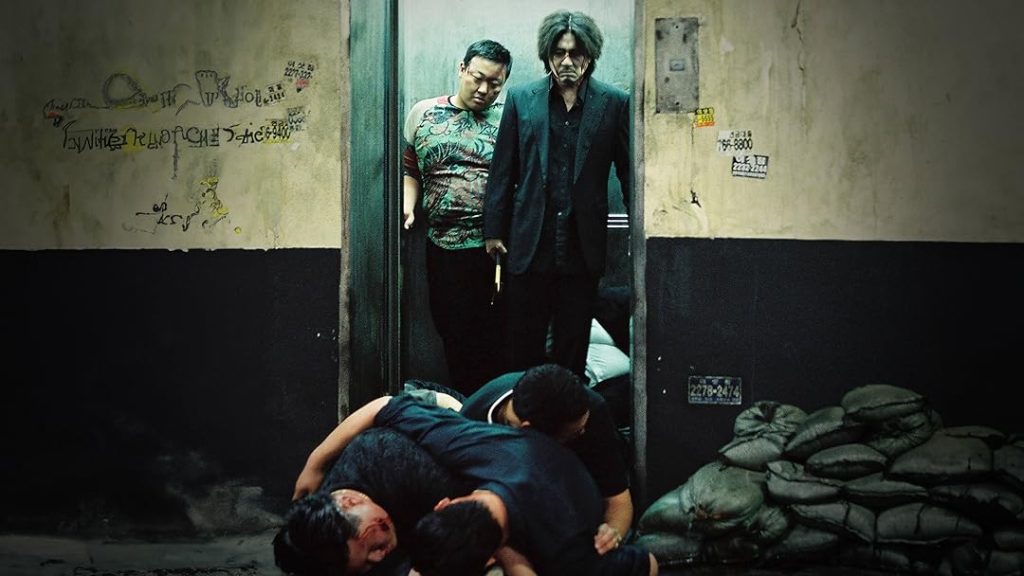 Ein Mann mit wildem Haar und Hammer steht im Aufzug über am Boden liegenden Männern in einem schmutzigen Flur.