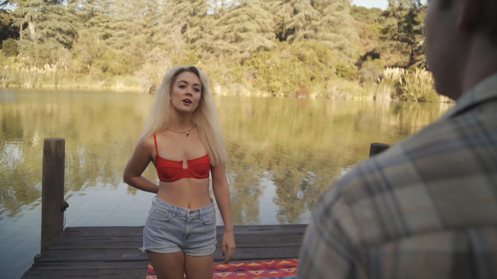 Eine junge blonde Frau mit einem roten Bikini-Oberteil und kurzen Jeans-Shorts steht auf einem Holzsteg am Ufer eines Sees. Sie schaut mit offenem Mund in Richtung eines Mannes, dessen Rückenansicht im Vordergrund zu sehen ist. Im Hintergrund sind Bäume und Schilf zu erkennen, die sich im sonnigen Seewasser spiegeln.