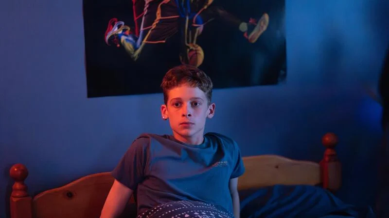 Ein jugendlicher Junge mit kurz geschnittenen, hellbraunen Haaren und großen Augen sitzt auf einem Holzrahmenbett. Er trägt ein einfaches blaues T-Shirt und eine gemusterte Pyjamahose. Sein Blick ist direkt auf den Betrachter gerichtet und wirkt überrascht oder schockiert. Im Hintergrund, an der blauen Wand, hängt ein Plakat, das einen Basketballspieler bei einem Dunking zeigt. Die Szene ist von einem starken, blauen Licht auf der linken Seite und einem rötlich-orangenen Licht auf der rechten Seite beleuchtet.
