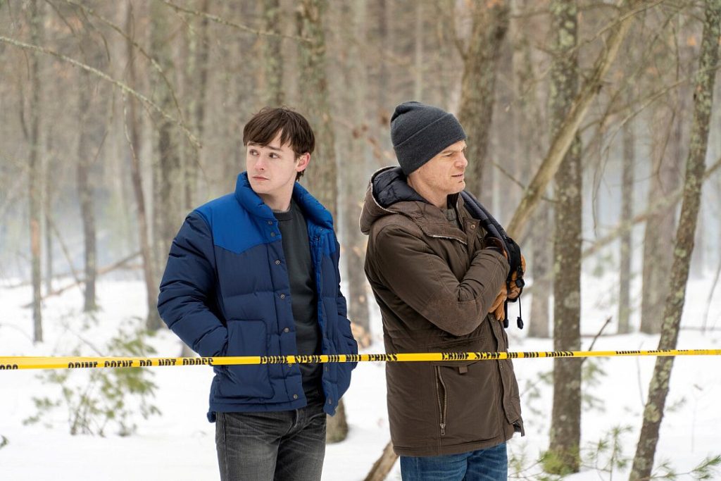 Zwei Männer, ein älterer und ein jüngerer, stehen in einem verschneiten Waldgebiet. Sie sind von gelbem Absperrband umgeben, das mit der Aufschrift „CRIME SCENE DO NOT CROSS“ versehen ist. Der ältere Mann trägt eine braune Jacke, eine graue Mütze und hat die Hände in den Taschen. Der jüngere Mann trägt eine blaue Steppjacke und blickt nachdenklich zur Seite. Beide scheinen in ein Gespräch vertieft zu sein oder etwas zu beobachten, das sich außerhalb des Bildes befindet. Im Hintergrund sind Bäume zu sehen.