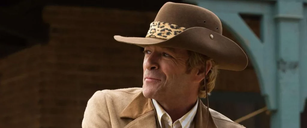 Nahaufnahme eines Mannes mittleren Alters, der einen braunen Cowboyhut mit einem Leopardenmuster-Band trägt. Er hat helle Haare, eine leicht runzlige Stirn und schaut mit einem leichten Lächeln nach links aus dem Bild. Er trägt eine beige Wildlederjacke über einem gestreiften Hemd. Im Hintergrund sind undeutlich braune und hellblaue Holzstrukturen zu erkennen.