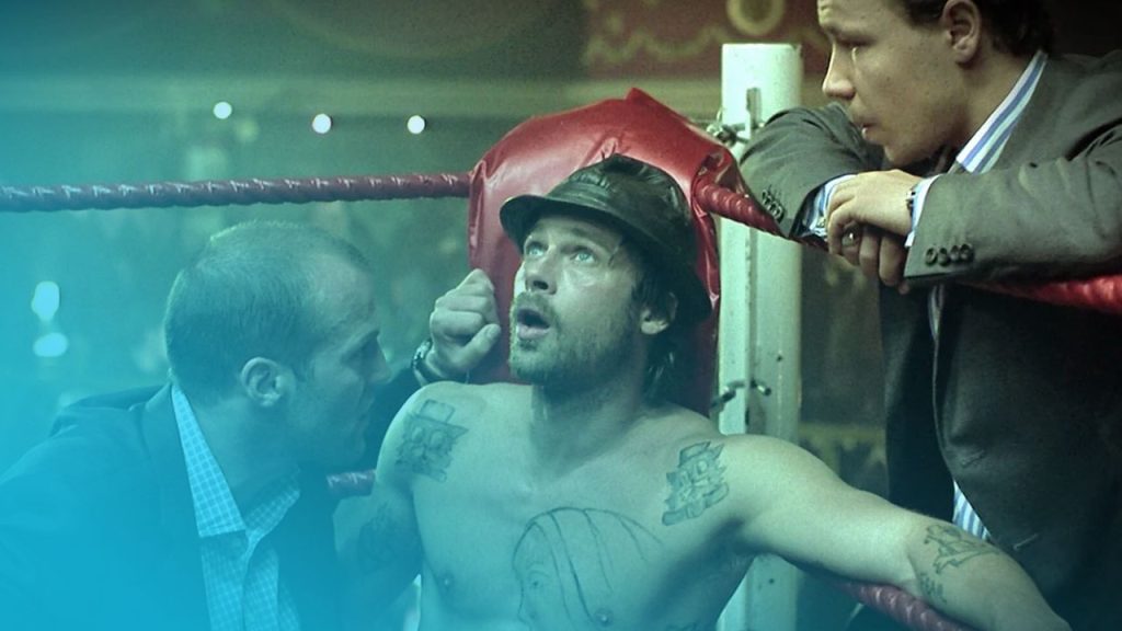 Ein Bild aus dem Film Snatch – Schweine und Diamanten, das drei Männer in einem Boxring zeigt. In der Mitte sitzt ein Mann mit freiem Oberkörper, tätowiert mit einem schreienden Gesicht auf der Brust und chinesischen Schriftzeichen auf den Schultern. Er trägt einen schwarzen Hut und blickt mit offenem Mund und einem erschrockenen oder verwirrten Gesichtsausdruck nach oben. Links von ihm steht ein Mann im dunklen Anzug mit einem karierten Hemd, der eine Hand auf die Schulter des sitzenden Mannes legt und zu ihm spricht. Rechts von ihm beugt sich ein weiterer Mann im grauen Anzug über die Boxringseile und schaut ebenfalls besorgt zu dem Mann im Hut. Der Hintergrund ist unscharf, man erkennt jedoch eine beleuchtete Sporthalle.