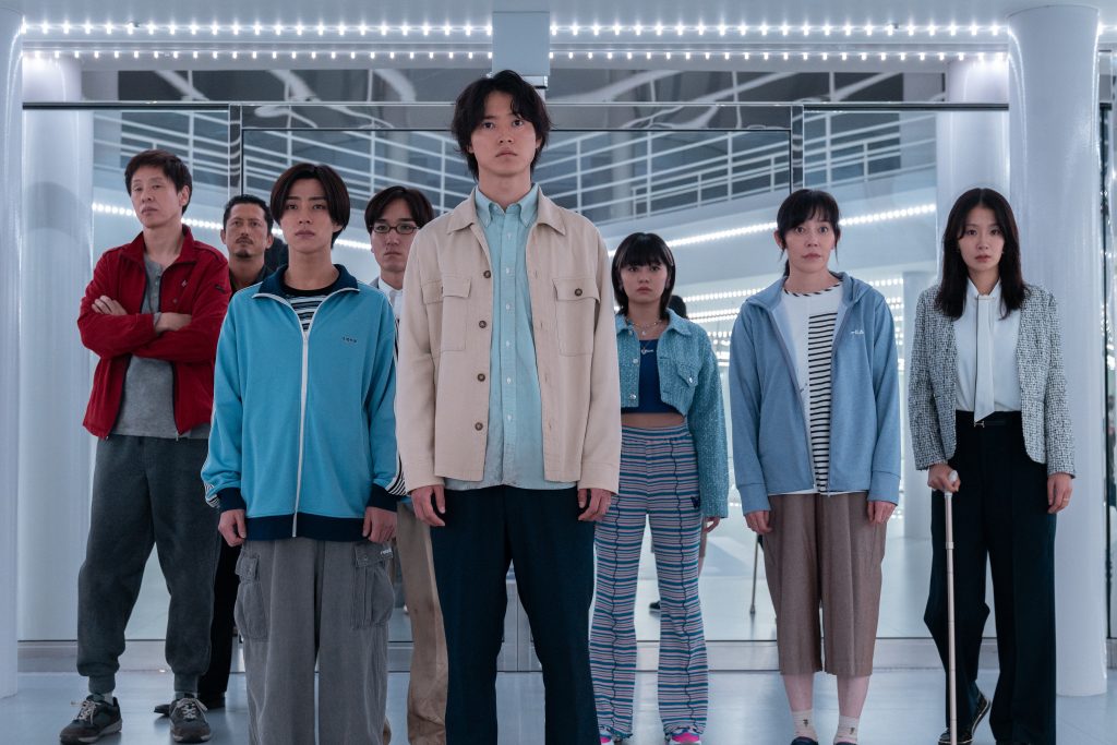 Eine Gruppe von acht Personen asiatischer Abstammung, die alle in die Kamera blicken, steht in einem hell erleuchteten, futuristisch wirkenden Raum mit spiegelnden Oberflächen und rechteckigen Lichtern an der Decke. Die Gruppe ist dicht gedrängt, mit einem großen Mann in der Mitte in einer beigen Jacke über einem hellblauen Hemd und dunklen Hosen. Links von ihm steht eine Person mit kurzen Haaren und einer hellblauen Sportjacke. Ganz links steht ein Mann in einer roten Jacke mit verschränkten Armen. Rechts vom Zentrum steht eine junge Frau in einer blauen, kurzen Jacke und gestreiften Hosen. Ganz rechts stehen zwei Frauen, eine mit einem hellblauen und weißen Pullover und die andere in einer schwarz-weiß gemusterten Jacke über einer weißen Bluse und dunklen Hosen. Alle Gesichter wirken ernst oder neutral.