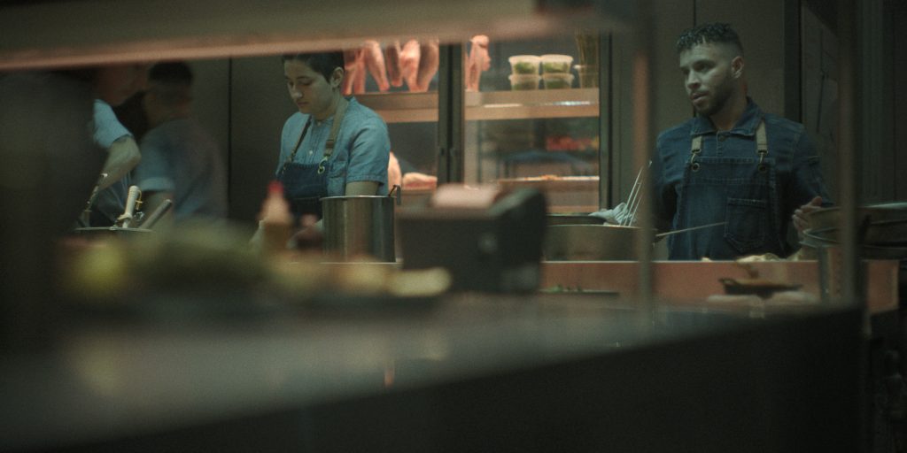 Eine verschwommene, dunkle Nahaufnahme einer belebten Restaurantküche. Im Hintergrund stehen zwei Köche, die an einer Theke arbeiten. Der Koch auf der rechten Seite, ein Mann mit kurzen, lockigen Haaren, trägt eine Jeansjacke und eine blaue Schürze und blickt zur Seite, während er etwas in einer Metallschüssel zubereitet. Die Köchin links neben ihm hat dunkle Haare und trägt ebenfalls eine blaue Schürze; sie blickt mit konzentriertem Gesichtsausdruck auf die Arbeitsfläche vor sich. Im Hintergrund, in einem Kühlraum hinter den Köchen, sind Fleischstücke zu sehen. Der Vordergrund ist verschwommen und zeigt eine reflektierende Theke mit verschwommenen Objekten, die darauf stehen. Das Bild hat einen graublauen Farbton.