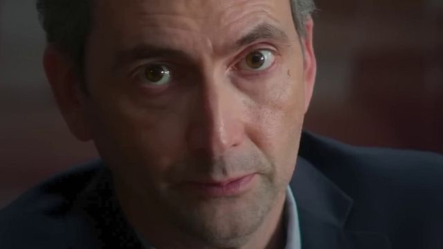 Nahaufnahme des britischen Schauspielers David Tennant. Er blickt direkt in die Kamera, seine Augen sind weit geöffnet und braun. Er hat dunkle Haare und auf seiner rechten Wange ist ein Muttermal zu sehen. Er trägt ein dunkelblaues Sakko und ein helles Hemd. Der Hintergrund ist unscharf und in warmen, rötlichen Tönen gehalten. Die Beleuchtung ist hell und von vorne.