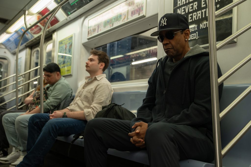 Ein Mann mittleren Alters, der eine New York Yankees-Kappe und eine Sonnenbrille trägt, sitzt auf einer blauen Bank in einer U-Bahn. Er trägt einen dunklen Kapuzenpullover und Hosen und hat die Hände im Schoß gefaltet. Neben ihm sitzt ein jüngerer Mann in einem hellbeigen Hemd. Im Hintergrund sind weitere Passagiere zu sehen, und an der Wand hängt ein Schild. Das Licht ist grell und reflektiert auf den Fenstern und den Oberflächen der U-Bahn.