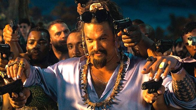 Ein Nahaufnahmebild des Schauspielers Jason Momoa in der Rolle des Dante Reyes aus dem Film "Fast X". Er hat schulterlanges braunes Haar, einen Oberlippenbart und Ziegenbart und trägt eine Sonnenbrille auf dem Kopf. Er trägt eine hellviolette, seidenähnliche Hemdjacke, eine Kette mit zahnähnlichen Anhängern und viele Ringe an seinen Fingern. In jeder Hand hält er eine schwarze Pistole und zielt damit direkt in die Kamera. Um ihn herum sind mehrere andere Personen zu sehen, die ebenfalls Pistolen in die Luft halten. Der Hintergrund ist unscharf und dunkel.