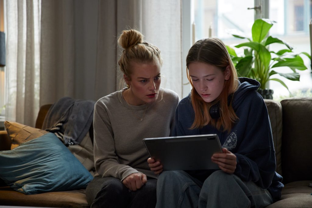 Zwei Frauen sitzen auf einem bequemen, braunen Sofa und schauen konzentriert auf ein Tablet, das sie gemeinsam halten. Die Frau links hat blonde Haare, die zu einem lockeren Dutt hochgesteckt sind. Sie trägt einen grauen Pullover und eine dunkle Hose. Ihr Gesichtsausdruck ist ernst und besorgt, mit leicht geöffnetem Mund und gerunzelter Stirn, während sie auf den Bildschirm blickt. Die jüngere Frau rechts, mit langen, glatten, braunen Haaren, trägt einen dunkelblauen Kapuzenpullover. Sie hat Kopfhörer um den Hals hängen und ihr Blick ist ebenfalls gespannt und auf das Tablet gerichtet. Das Licht fällt von einem großen Fenster hinter ihnen in den Raum, wo auch eine grüne Topfpflanze steht.