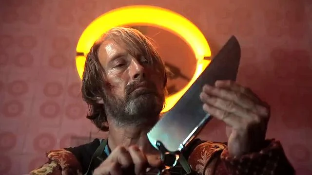 Ein Mann mit zerzaustem Haar und Bart, der blutige Kratzer im Gesicht hat, hält ein großes, spiegelndes Küchenmesser vor sich und blickt nachdenklich auf die Klinge. Im Hintergrund ist eine leuchtend orangefarbene, ringförmige Leuchte zu sehen, die an einer rosafarbenen Wand mit einem kreisförmigen Muster hängt. Der Mann trägt eine Weste über einem blauen Hemd.