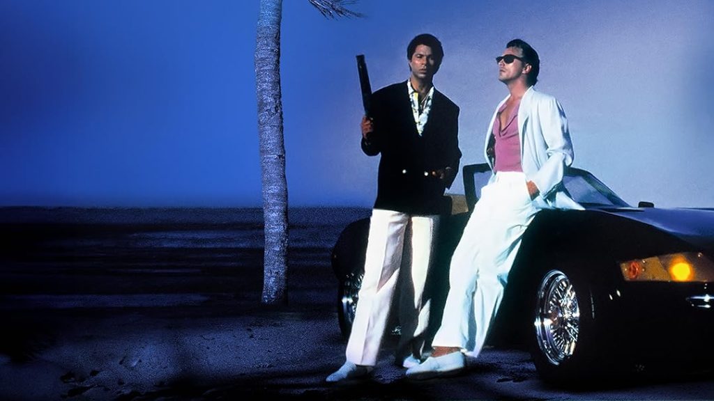 Zwei Männer im Stil der 80er Jahre stehen nachts auf einem dunklen Strand vor einem schwarzen Cabriolet. Der linke Mann trägt eine schwarze Jacke über einem hellen Hemd, eine weiße Hose und hält einen schwarzen Gegenstand (möglicherweise eine Waffe) über der Schulter. Der rechte Mann lehnt an der Motorhaube des Autos und trägt einen weißen Anzug mit offenem Jackett, ein pinkes T-Shirt und eine Sonnenbrille. Links ist der dunkle Stamm einer Palme zu sehen. Die Szene ist in dunkles, tiefes Blau getaucht, was eine geheimnisvolle, nächtliche Stimmung erzeugt.