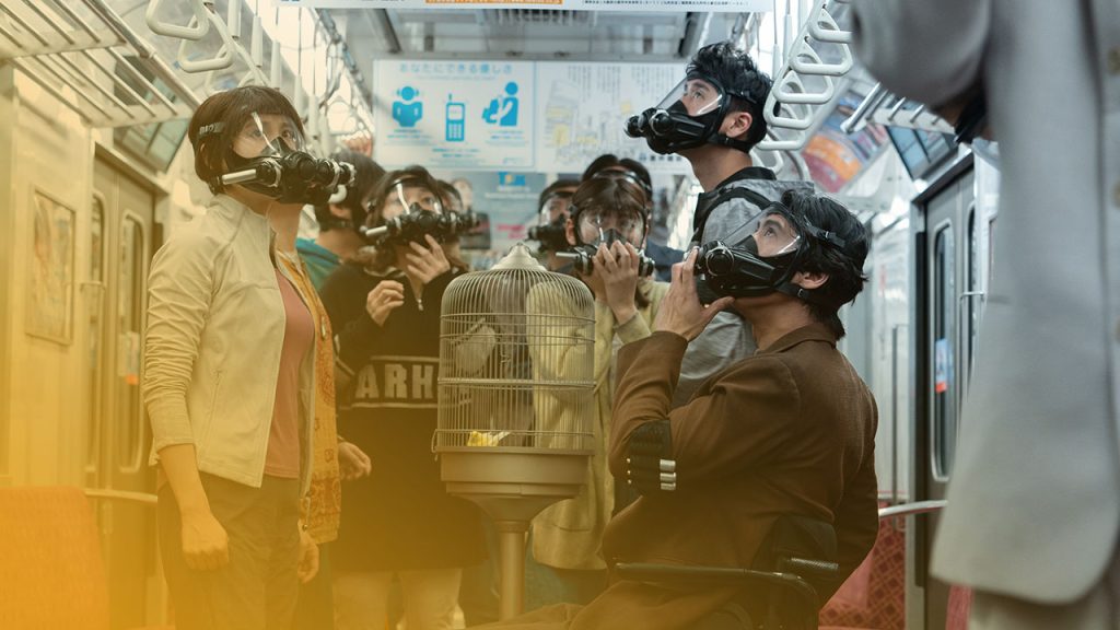 Eine Nahaufnahme in einem U-Bahn-Waggon zeigt eine Gruppe von etwa zehn Personen, die alle schwarze Gasmasken mit klaren Sichtfenstern tragen, die ihre Nasen und Münder bedecken. Im Vordergrund sitzt ein Mann in einem braunen Sakko auf einem Rollstuhl und blickt nach oben und rechts. Neben ihm steht eine Frau in einer hellen Jacke und grauen Hose, die ebenfalls eine Gasmaske trägt. In der Mitte des Waggons steht ein großer, runder Vogelkäfig auf einem Ständer, in dem sich vermutlich ein Vogel befindet. Die anderen maskierten Personen stehen hinter dem Käfig und schauen ebenfalls nach oben oder auf den Käfig. Die Sitze des Zuges sind mit einem gemusterten, magenta-roten Stoff bezogen. Über den Köpfen befinden sich japanische Werbung und Hinweisschilder. Die gesamte Szene wirkt angespannt und beunruhigend.