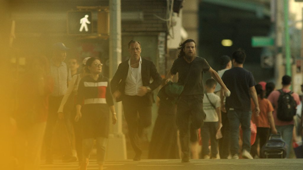 Zwei Männer rennen in einer belebten Stadt auf der Straße, an Fußgängern vorbei. Der Mann vorne hat einen Bart, dunkle, zerzauste Haare und trägt eine grüne Tasche und ein schwarzes T-Shirt. Der Mann hinter ihm ist glattrasiert, trägt ein schwarzes Sakko, ein weißes Hemd und eine schwarze Hose. Ein Fußgängerüberweg und ein weißes Fußgängerzeichen an einer Ampel sind im Bild zu sehen. Die Szene ist hell und leicht neblig, was auf späten Nachmittag hindeutet.