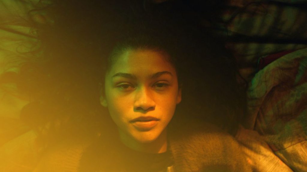 Eine Nahaufnahme von Zendaya, die flach auf dem Rücken liegt und direkt in die Kamera blickt. Ihr langes, dunkles Haar breitet sich um ihren Kopf aus. Ihr Gesicht wird von einem starken, warmen gelben Licht von unten und einem kalten, grünen Licht von oben beleuchtet. Sie trägt einen dunklen Pullover und ihr Ausdruck ist neutral, aber mit weit geöffneten Augen.