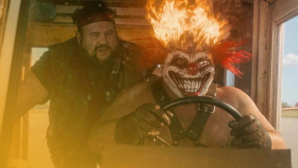 Ein großer, kräftiger Mann mit Bart und rotem Stirnband steht im Hintergrund und blickt zur Seite. Im Vordergrund sitzt eine männliche Figur mit einer bedrohlich grinsenden Clownsmaske mit roter Nase und großen Zähnen, roten und gelben Haaren, die an den Spitzen brennen, und trägt einen schwarzen Ledergurt und Handschuhe. Die Figur hält das Lenkrad eines Fahrzeugs in einem hell erleuchteten Innenraum.