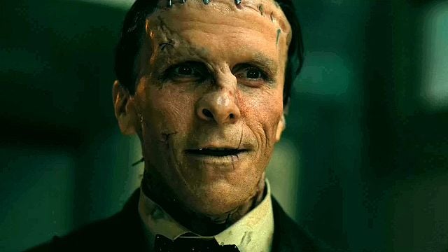 Nahaufnahme des Schauspielers Christian Bale als Frankensteins Monster, der mit Narben, unebener Haut und Stichen im Gesicht und an der Stirn zu sehen ist. Er trägt einen schwarzen Anzug mit Fliege, ein beiges Hemd und eine Weste. Er lächelt leicht und schaut nach vorne, während er sich in einem dunklen, unordentlichen Raum mit einer Lampe und einem Tisch befindet.