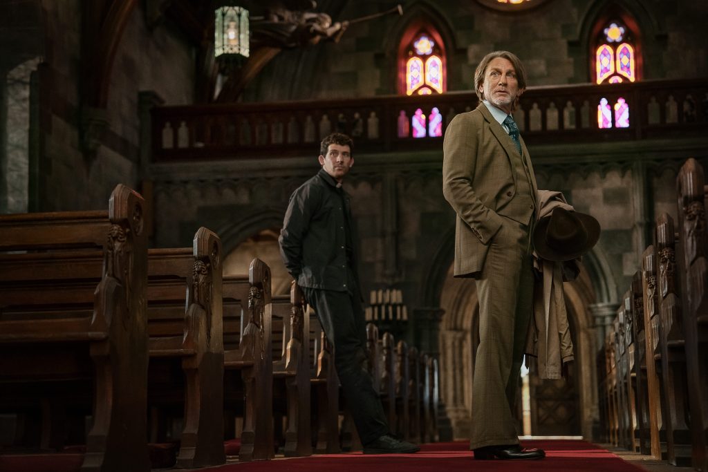 Zwei Männer in einer alten, dunklen Kirche. Ein älterer Mann in einem braunen Tweed-Anzug und mit einem Hut in der Hand steht im Vordergrund. Sein Blick ist nachdenklich oder besorgt. Hinter ihm, im Schatten, lehnt ein jüngerer Mann in einer schwarzen Jacke an einer Kirchenbank und blickt zur Seite. Der Boden ist mit einem roten Teppich ausgelegt, der durch die Gänge zwischen den Holzpänken führt. Buntes Licht fällt durch gotische Bogenfenster. Im Hintergrund ist eine Empore und eine große Skulptur an der Decke zu erkennen.