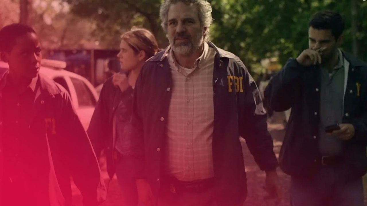 Eine Gruppe von vier FBI-Agenten, gekleidet in blaue Jacken mit der gelben Aufschrift "FBI", geht auf einem schlammigen Feldweg. Die Gruppe besteht aus einem Mann mit lockigem graumeliertem Haar in der Mitte, der den Betrachter ansieht, einem Afroamerikaner zu seiner Linken, einer Frau hinter ihm und einem Mann zu seiner Rechten, der in sein Handy blickt. Im Hintergrund sind Bäume, ein Polizeiauto und weitere Personen zu sehen. Das Bild hat einen düsteren, ernsten Ton.