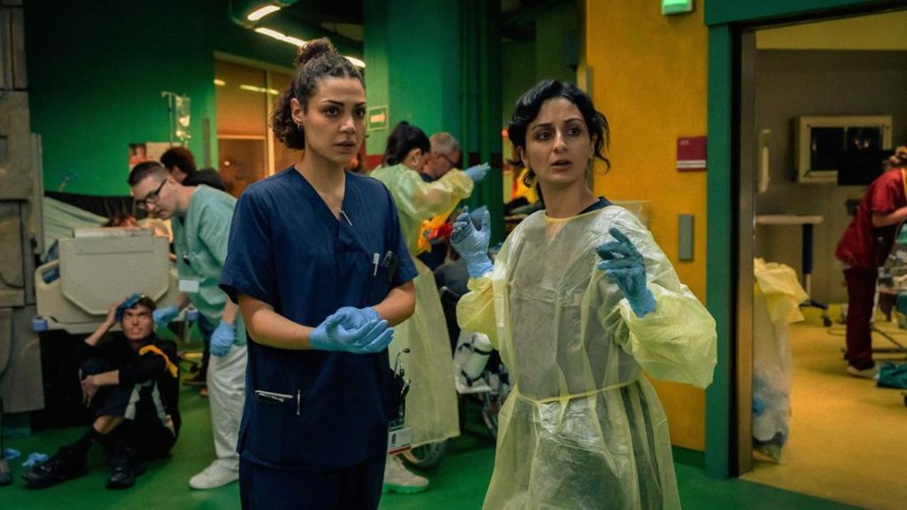 Ein Szenebild in einem überfüllten Krankenhausflur. Im Vordergrund stehen zwei Frauen: Die linke trägt blaue Krankenhauskleidung und blaue Handschuhe, blickt nach rechts und hat die Hände nach vorne gestreckt. Ihre Haare sind zu einem unordentlichen Dutt hochgesteckt. Die Frau rechts trägt einen gelben, durchsichtigen Schutzkittel über ihrer Kleidung, blaue Handschuhe und hat ihre Hände in die Luft gehoben, als ob sie gestikulieren würde. Ihr Gesichtsausdruck ist besorgt. Sie hat lockiges, dunkles Haar, das bis zu den Schultern reicht. Im Hintergrund sind weitere Personen in medizinischer Kleidung zu sehen, darunter ein Mann, der verängstigt auf dem Boden sitzt. Die Wände sind in leuchtendem Gelb und Grün gestrichen und das Bild ist von chaotischer und angespannter Stimmung geprägt.