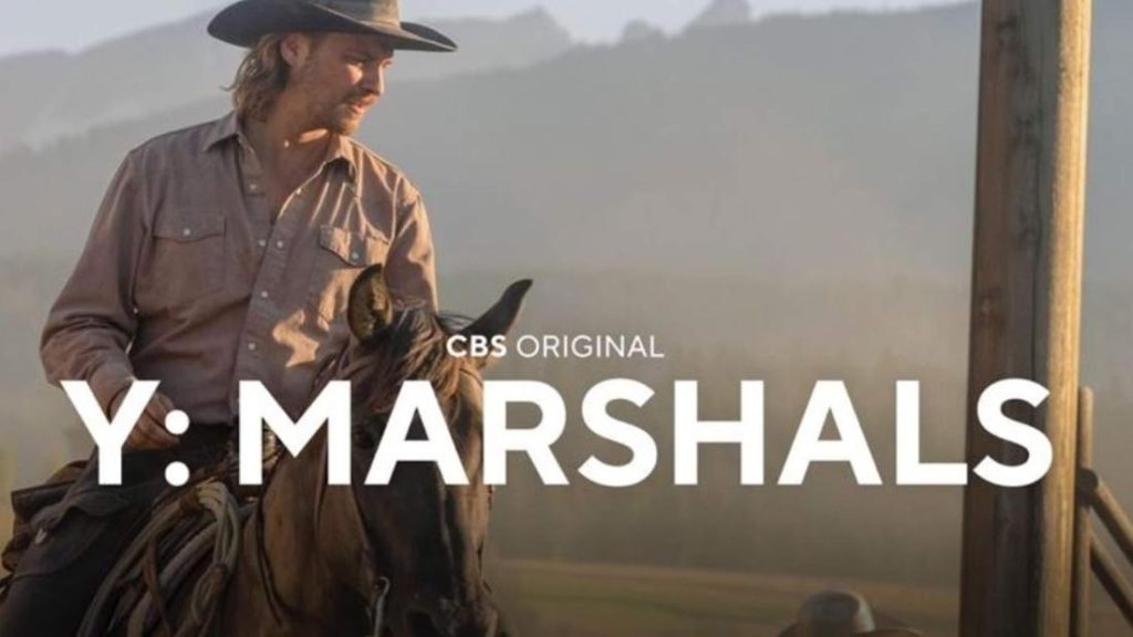 Nahaufnahme von Luke Grimes als Cowboy, der mit einer hellbraunen Bluse und einem dunklen Cowboyhut auf einem braunen Pferd reitet. Er blickt nach rechts. Der Hintergrund ist eine verschwommene, helle Landschaft mit bergähnlichen Umrissen. Über dem Bild liegt ein großer, weißer Text der Serie oder des Films "Y: MARSHALS" mit dem kleineren Text "CBS ORIGINAL" darüber. Die Beleuchtung ist weich und diffus.