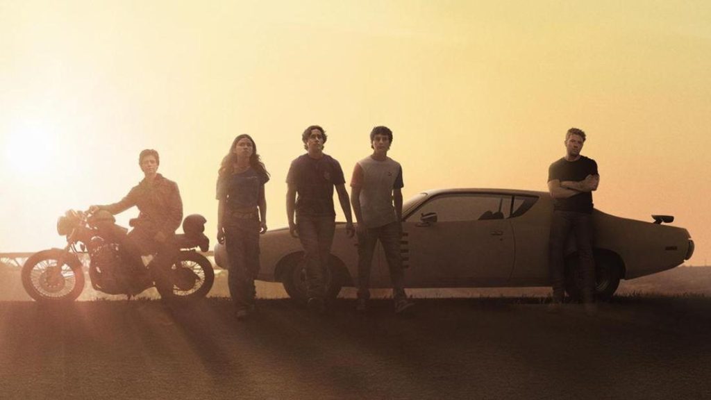 Gruppenfoto von fünf Personen und zwei Fahrzeugen bei Sonnenuntergang. Von links nach rechts: Eine Person sitzt auf einem Motorrad. Eine junge Frau mit langen Haaren geht, gefolgt von zwei jungen Männern. Rechts steht ein älterer Mann mit verschränkten Armen lässig an einem klassischen Muscle-Car, das die Mitte und den rechten Teil des Bildes dominiert. Der Horizont ist tief, die Sonne steht sehr tief. Die Figuren sind vor dem hellen, ockerfarbenen Himmel in Silhouette zu sehen.