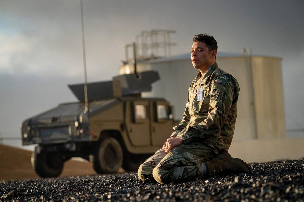 Ein männlicher Soldat in Tarnuniform kniet auf einem Kiesboden in einer Außenaufnahme. Er blickt nach rechts oben, wobei sein Gesicht leicht angestrahlt wird. Im Hintergrund, unscharf, ist ein militärisches Geländefahrzeug, wahrscheinlich ein Humvee, und dahinter eine große, helle Struktur, möglicherweise ein Tank oder ein Gebäude, zu sehen. Die Beleuchtung ist dramatisch mit einem dunklen, bewölkten oder dämmrigen Himmel.