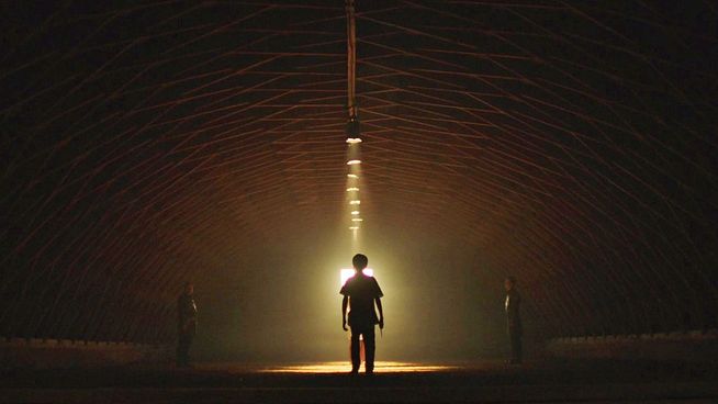 Ein einzelner Mann steht als Silhouette in der Mitte einer kuppelartigen oder gewölbten Struktur, beleuchtet von einem hellen, herabscheinenden Lichtstrahl, der direkt über ihm von der Decke hängt und ihn in eine leuchtend gelbe oder goldene Aura taucht. Zwei weitere Figuren sind nur schemenhaft als Silhouetten weiter außen links und rechts von ihm im Halbdunkel zu erkennen. Die gesamte Szene ist in dunklen Braun- und Schwarztönen gehalten, mit dem hellen Gegenlicht, das die Personen dramatisch hervorhebt.