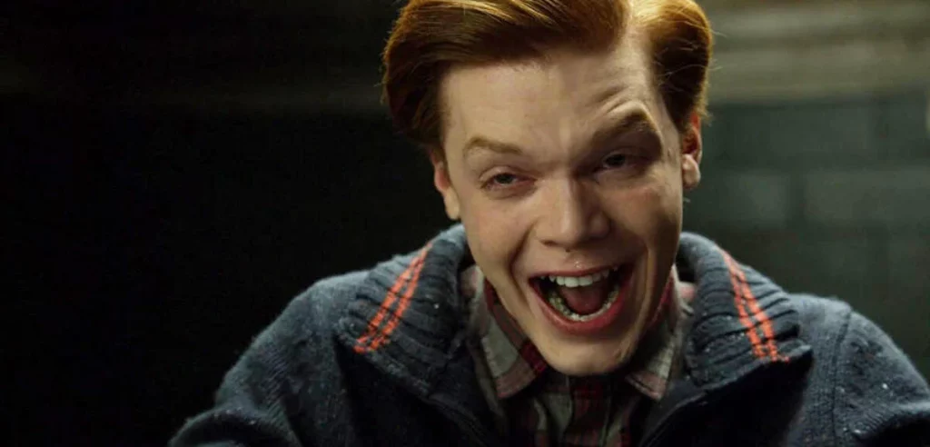 Nahaufnahme des Schauspielers Cameron Monaghan in seiner Rolle als Jerome Valeska oder Jeremiah Valeska aus der Serie *Gotham*. Er hat leuchtend rotes Haar und trägt einen dunkelblauen, gestrickten Cardigan mit orangefarbenen und weißen Streifen an der Schulter und Kragen. Er blickt direkt in die Kamera mit aufgerissenem Mund und einem manischen, breiten Grinsen, wobei seine Augen leicht schielen. Der Hintergrund ist sehr dunkel und unscharf.