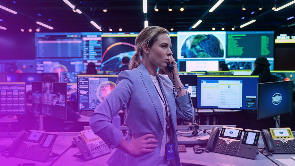 Eine blonde Frau mittleren Alters in einem hellblauen Anzug steht in einem hochtechnologischen Kontrollraum, den Hörer eines Telefons an ihr rechtes Ohr haltend. Ihre linke Hand stützt sie in die Hüfte. Der Raum ist dunkel, aber beleuchtet von zahlreichen Monitoren und Bildschirmen im Hintergrund, die verschiedene Karten, Daten und Grafiken anzeigen, einschließlich einer Darstellung der Erde. Mehrere Festnetztelefone und Tastaturen liegen auf grauen Schreibtischen im Vordergrund. Andere Personen sind verschwommen im Hintergrund bei ihrer Arbeit zu sehen. Die Atmosphäre ist angespannt und beschäftigt.