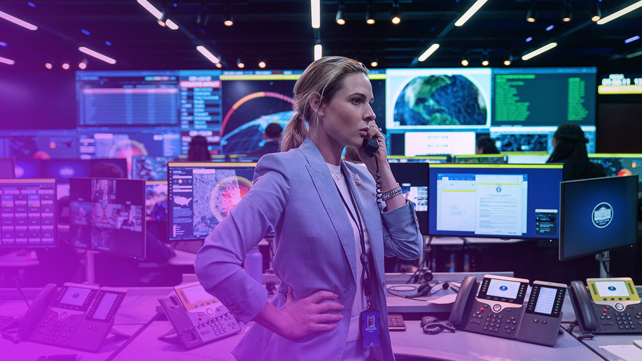 Eine blonde Frau mittleren Alters in einem hellblauen Anzug steht in einem hochtechnologischen Kontrollraum, den Hörer eines Telefons an ihr rechtes Ohr haltend. Ihre linke Hand stützt sie in die Hüfte. Der Raum ist dunkel, aber beleuchtet von zahlreichen Monitoren und Bildschirmen im Hintergrund, die verschiedene Karten, Daten und Grafiken anzeigen, einschließlich einer Darstellung der Erde. Mehrere Festnetztelefone und Tastaturen liegen auf grauen Schreibtischen im Vordergrund. Andere Personen sind verschwommen im Hintergrund bei ihrer Arbeit zu sehen. Die Atmosphäre ist angespannt und beschäftigt.