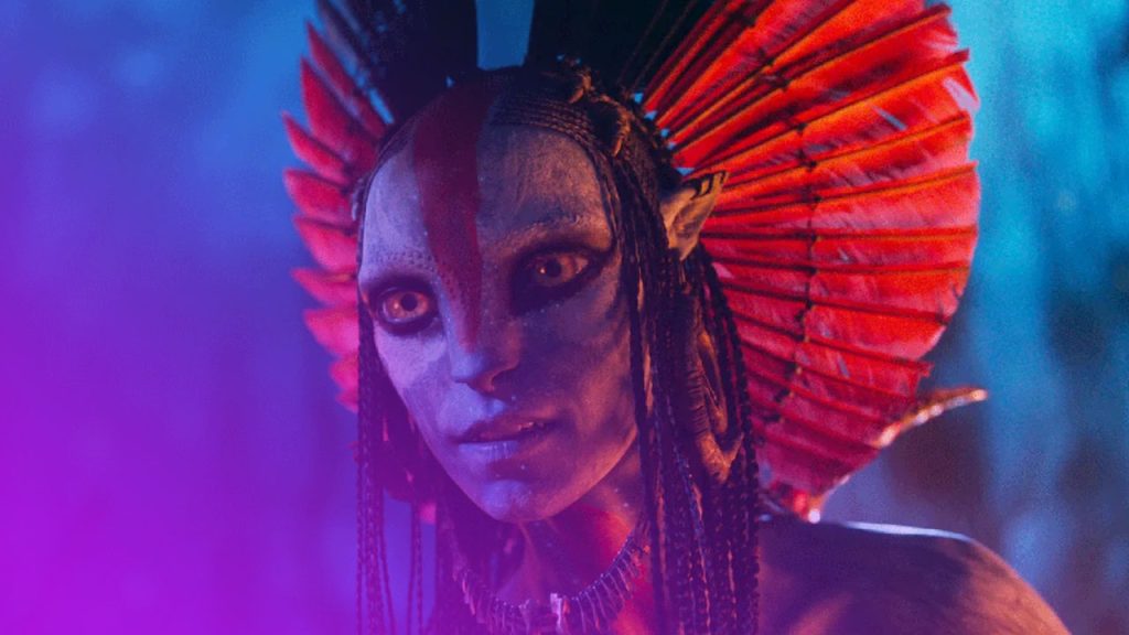 Nahaufnahme des Oberkörpers eines Na'vi, dessen blaue Haut mit roten Stammesmarkierungen auf der Stirn und über die Nase bemalt ist. Die Figur blickt direkt nach vorne mit weit geöffneten, goldenen Augen und trägt einen großen Kopfschmuck aus leuchtend roten Federn oder fächerartigen Elementen, die hinter ihrem Kopf aufgefächert sind. Mehrere schwarze Zöpfe rahmen das Gesicht ein. Der Hintergrund ist ein unscharfes, dunkles Blau, was eine mystische, nächtliche Stimmung erzeugt.