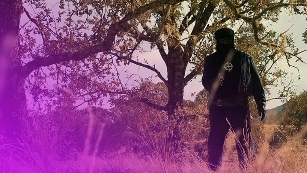 Ein düster wirkender Mann in komplett schwarzer Kleidung steht mit dem Rücken zum Betrachter in einer trockenen, grasbewachsenen Landschaft. Er trägt eine schwarze Gesichtsbedeckung oder Sturmhaube und auf dem Rücken seiner schwarzen Jacke ist ein weißer Kreis mit einem Fadenkreuz-Symbol zu sehen. Um ihn herum befinden sich hohe, trockene Gräser im Vordergrund und Bäume mit spärlichem Laub und viel Geäst. Die Szene ist von einem leicht gelblichen, natürlichen Licht durchflutet, was der Umgebung eine herbstliche oder trockene Stimmung verleiht.