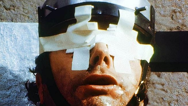 Ein dramatisches Nahaufnahme-Porträt eines Mannes, dessen Augen mit weißen Klebestreifen in Form von Kreuzen bedeckt sind. Er trägt einen dunklen, metallischen Helm oder eine Kappe, die mit großen Nägeln durchschlagen zu sein scheint, und sein Gesicht ist verschwitzt oder ölig. Der Hintergrund ist eine raue, beige-braune Oberfläche, möglicherweise eine Mauer. Die Beleuchtung ist hell und intensiv, was Schatten und eine rohe, unheimliche Stimmung erzeugt. Das Bild hat einen körnigen, filmischen Look.