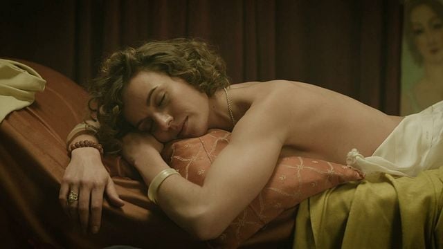 Eine hellhäutige Person mit kurzen, lockigen, braunen Haaren liegt mit geschlossenen Augen auf einem Bett und schläft. Sie liegt auf dem Bauch, den Kopf auf einem orangefarbenen Kissen, das von einem goldbraunen Kissen umgeben ist. Die Person ist an der Taille unbekleidet, der obere Rücken ist sichtbar. Sie trägt eine dünne Goldkette, einen Ring an der linken Hand, ein Armband aus rötlichen Perlen am linken Handgelenk und ein dickes, helles Armband (möglicherweise Jade) am rechten. Im Hintergrund rechts hängt an einer dunklen Wand ein Gemälde, das undeutlich eine Figur abbildet. Die Beleuchtung ist gedämpft und warm.