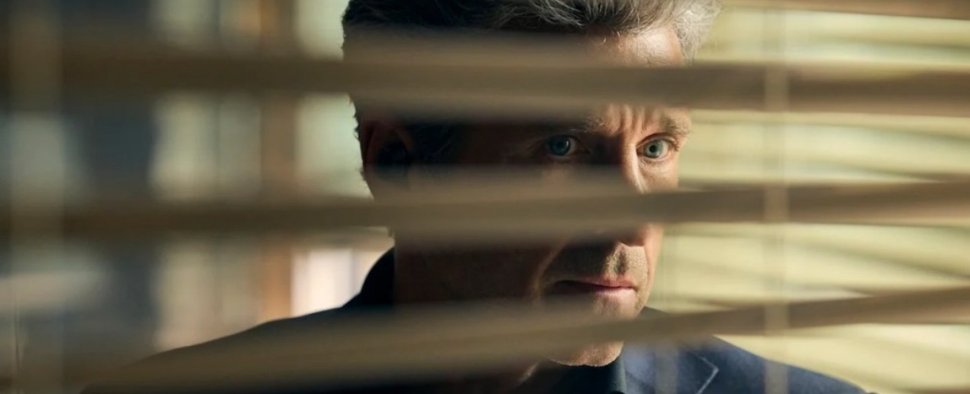 Ein dramatischer Nahaufnahme-Screenshot eines Mannes mit graumeliertem Haar und intensiven blauen Augen, der besorgt oder konzentriert durch die Lamellen einer horizontalen Jalousie blickt. Nur sein oberer Teil des Gesichts und die Augen sind zwischen den Lamellen sichtbar. Die Beleuchtung ist dunkel und schattenreich, was die gespannte Stimmung verstärkt. Er trägt ein dunkles Sakko oder Hemd.