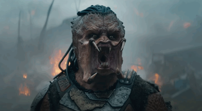 Nahaufnahme des Kopfes und Oberkörpers eines Predators mit rötlich-brauner, runzeliger Haut, geöffnetem, bedrohlichem Maul, das spitze Zähne und Reißzähne (Mandibeln) freilegt, und dunklen, geflochtenen Dreadlocks. Er trägt eine dunkle, zerfetzte Rüstung über der Schulter und der Brust. Der Hintergrund ist verschwommen und düster mit Rauch und orange-gelben Feuerschein von Flammen, was eine Atmosphäre von Kampf und Zerstörung vermittelt. Sein Ausdruck ist wütend oder aggressiv.