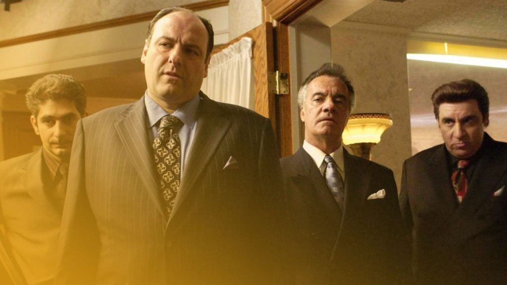 Ein Standbild mit vier Schauspielern aus der Serie Die Sopranos (The Sopranos) in Anzügen, die in einem Innenraum stehen. James Gandolfini steht in der Mitte und blickt nach vorne, gekleidet in einen dunkelblauen Nadelstreifenanzug mit einer gemusterten Krawatte. Links von ihm steht Michael Imperioli in einem dunklen Anzug mit violettem Hemd. Rechts von Gandolfini stehen Frank Vincent und Steven Van Zandt, beide in dunklen Anzügen und Krawatten. Der Hintergrund ist leicht verschwommen, zeigt einen Türrahmen mit einem weißen Vorhang und eine Ecke eines Raumes mit einem gelblich beleuchteten Bereich. Die Beleuchtung ist insgesamt warm und leicht düster.