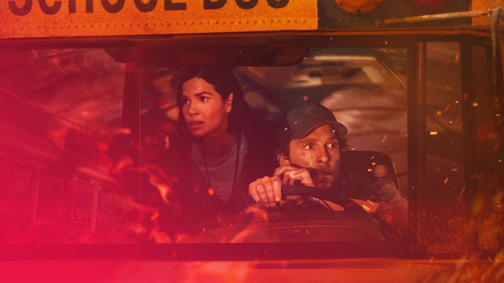 Ein mittelnaher Schuss durch die Windschutzscheibe eines Schulbusses zeigt einen Mann am Steuer und eine Frau daneben, beide blicken mit besorgtem Ausdruck nach vorne. Das Innere und Äußere des Busses, einschließlich der unteren Hälfte der Windschutzscheibe, sind von einem hellen, orange-roten Schein und Flammen umgeben, was auf einen Brand oder eine Explosion hindeutet. Der Mann trägt eine Baseballkappe und hält das Lenkrad fest. Die Frau ist leicht über seine Schulter gebeugt. Im oberen Teil des Bildes ist der gelbe Schriftzug "SCHOOL BUS" zu erkennen. Die Szene ist von Hitze und Gefahr geprägt.