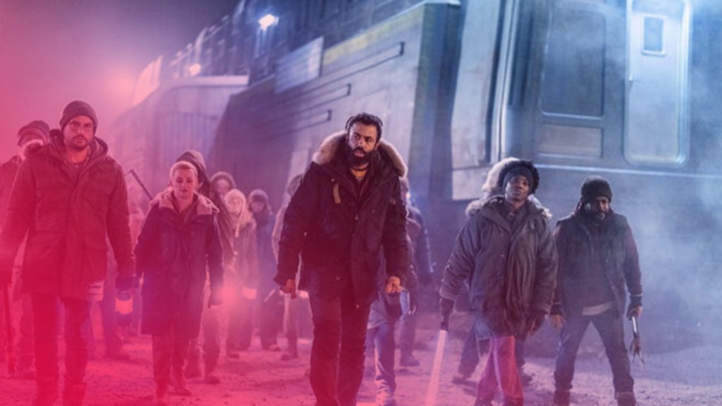 Eine Gruppe von etwa einem Dutzend Menschen in dicken Wintermänteln geht bei Nacht vor einem großen, hell beleuchteten Zug her. Im Vordergrund in der Mitte ist ein Mann mit einem dunklen Bart und einer schwarzen Kapuzenjacke zu sehen, der entschlossen blickt. Die anderen tragen ähnliche, gedämpfte Kleidung in Braun- und Grüntönen. Mehrere Personen halten kleine, blau leuchtende Lampen oder Stäbe. Die Szene ist von Schnee und Kälte geprägt und die Beleuchtung ist dramatisch.