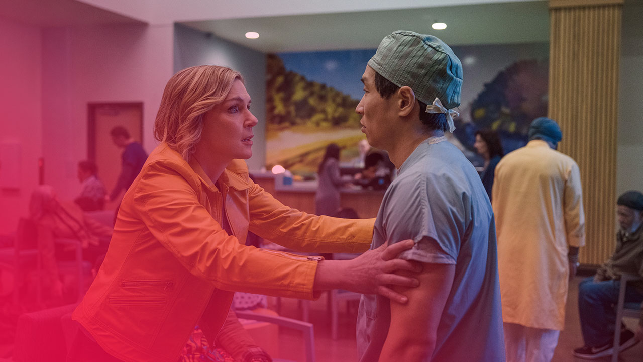 Eine blonde Frau in einer auffälligen, senfgelben Lederjacke spricht intensiv mit einem männlichen Arzt in türkisfarbenem OP-Kittel und Haube. Sie hält seine Arme und Schultern fest, beide haben ernste Ausdrücke in einem Krankenhaus-Wartebereich mit gedämpftem Licht. Im Hintergrund sind undeutlich weitere Personen und eine große Wandmalerei mit Bäumen zu sehen.