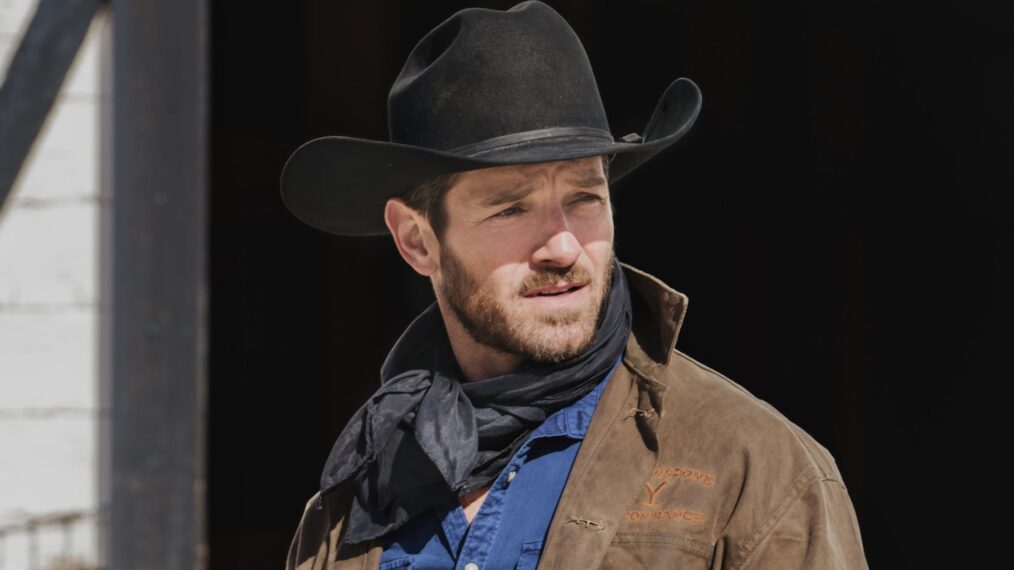 Brustbild eines bärtigen Mannes mit Cowboyhut und Halstuch. Er trägt einen schwarzen Cowboyhut, ein schwarzes Halstuch lose um den Hals, ein dunkelblaues Hemd und eine braune Jacke mit einem gelben Schriftzug auf der Brust. Er blickt nach rechts aus dem Bild. Der Hintergrund ist unscharf und dunkel, was den Mann hervorhebt.
