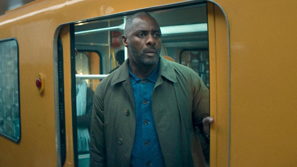 Ein Mann mit Bart, bekleidet mit einem blauen Hemd und einem olivgrünen Trenchcoat, steht in der offenen Tür eines U-Bahn- oder Zugwagons, der außen leuchtend gelb ist. Er blickt mit ernstem Ausdruck nach rechts aus dem Bild. Im Hintergrund des Wagens sind undeutlich weitere Personen und die Inneneinrichtung zu erkennen.