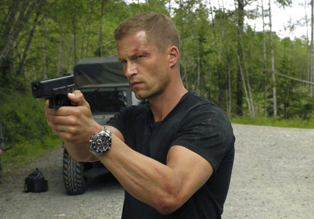 Mann mit kurzer, blonder Frisur zielt mit schwarzer Pistole. Er trägt ein schwarzes T-Shirt und eine silberne Armbanduhr. Im Hintergrund ein Militärfahrzeug und Bäume.