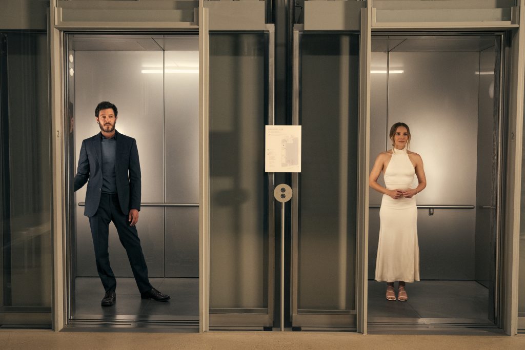 Ein Mann im Anzug und eine Frau im weißen, trägerlosen Kleid stehen in getrennten, leicht geöffneten Aufzügen.